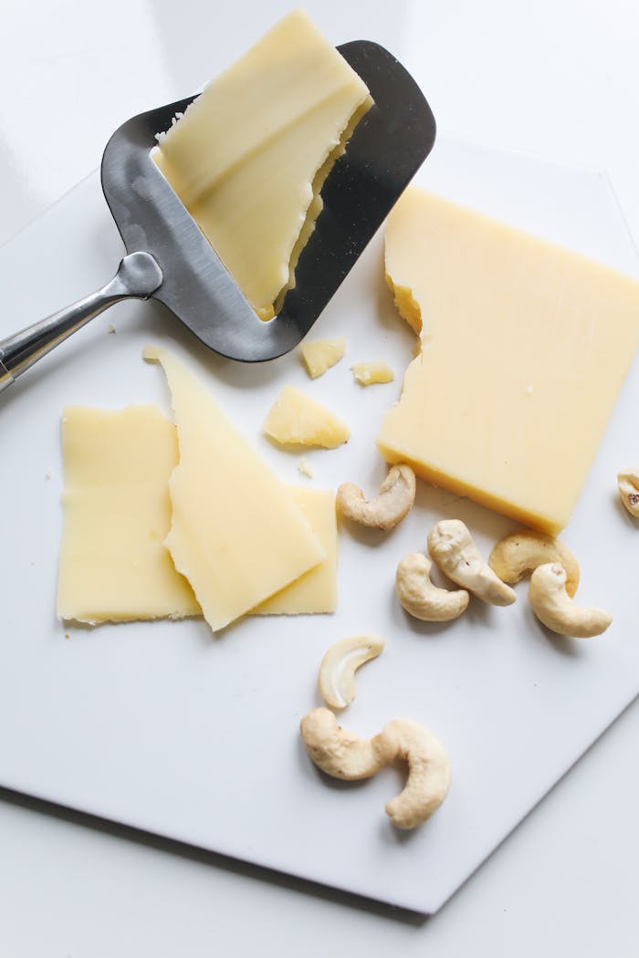 Sliced cheese and cashews on a white hexagonal board with a cheese slicer, minimal style.